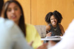 A person with curly hair is smiling while sitting at a table with a laptop and notebooks. They appear to be engaged in a meeting or discussion with others.