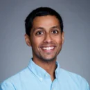 Headshot of a person smiling, wearing a light blue collared shirt, against a plain gray background.