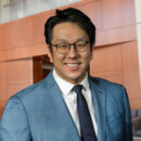 A person in a blue suit, white shirt, and black tie, smiling, wearing glasses. They are standing in a modern office environment with wooden panels and contemporary furnishings. This is a headshot.