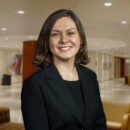 A person in a professional headshot wearing a dark blazer, smiling, standing in a well-lit modern office or lobby area.