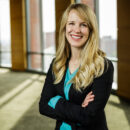 A person with long blond hair, wearing a black blazer and blue top, smiles confidently with arms crossed in a sunlit indoor setting with large windows. This represents a professional or business context.