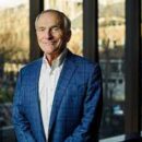 A smiling person in a blue checkered suit jacket and white shirt stands in front of a window with a blurred outdoor background.