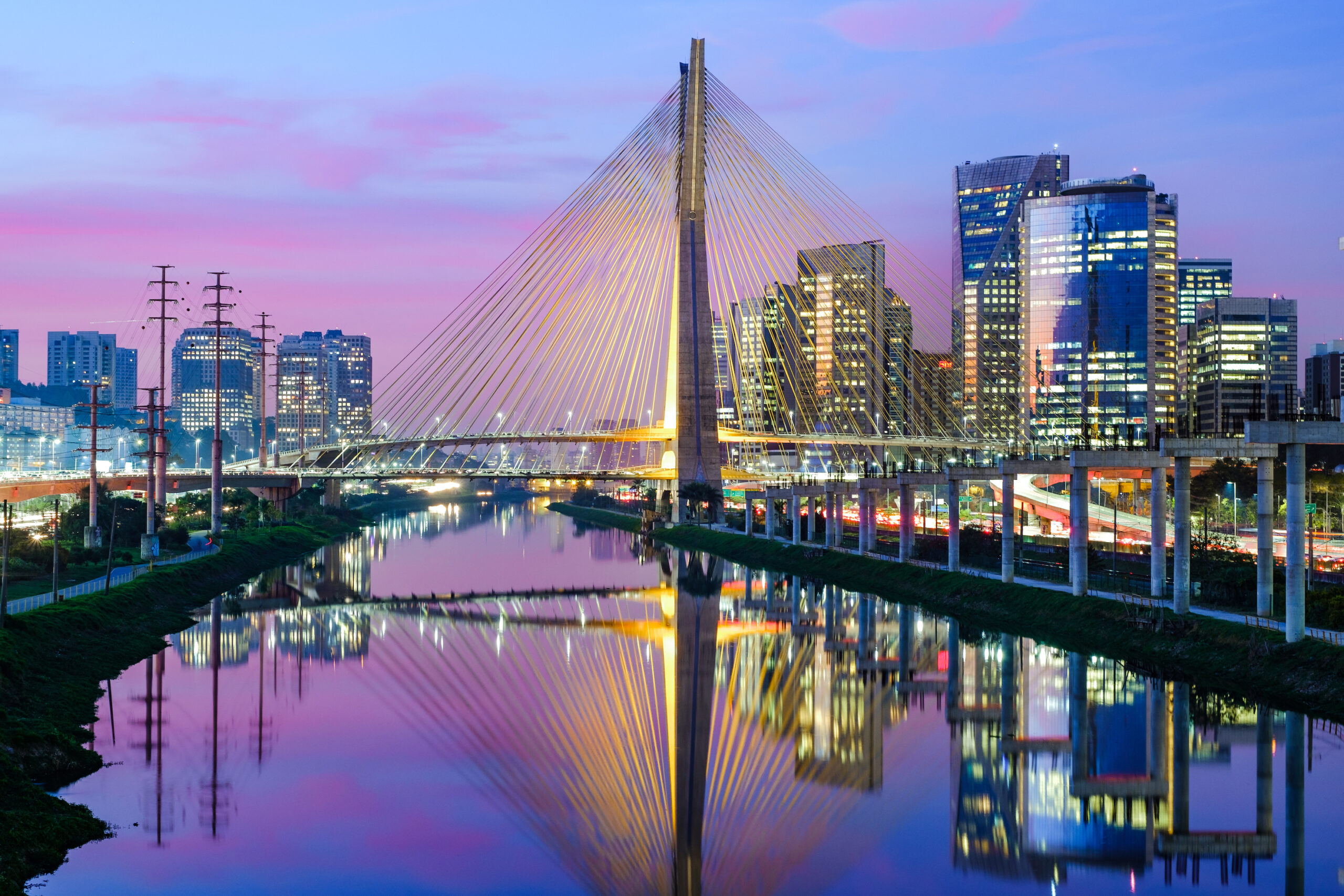 The image depicts the Octávio Frias de Oliveira Bridge in São Paulo at twilight, with illuminated skyscrapers and a colorful sky reflecting in the river below.