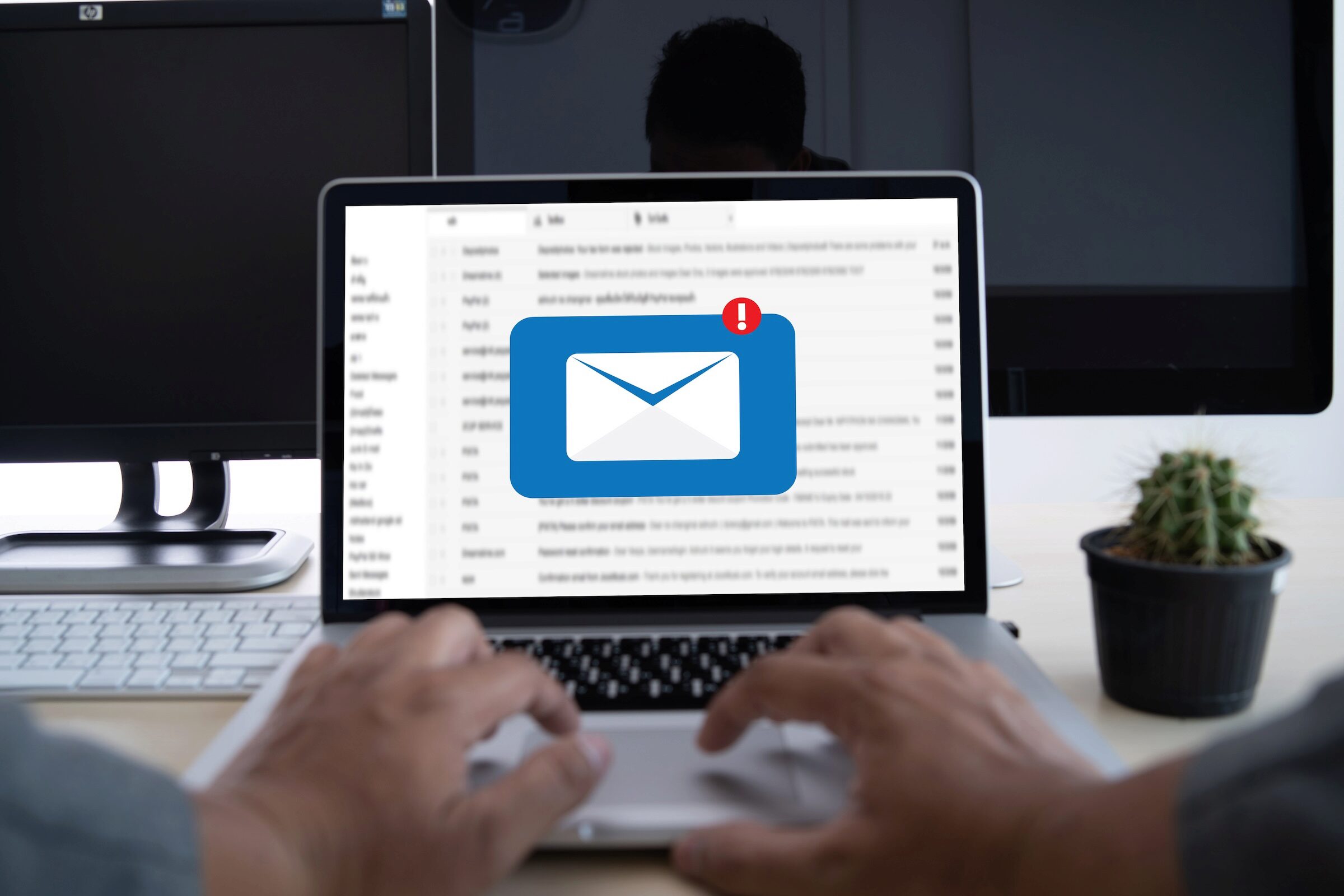 Person typing on a laptop displaying an email inbox with a notification icon. A potted cactus sits nearby on the desk.