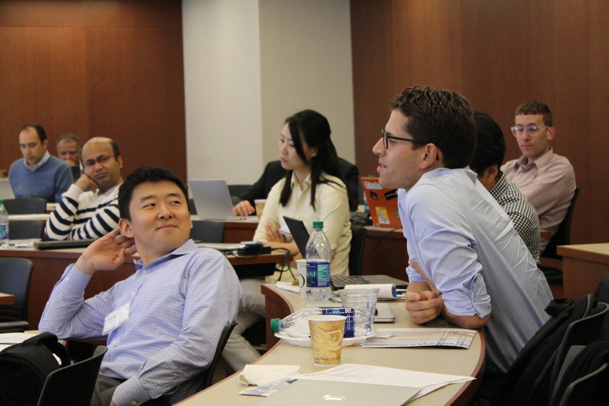 A small group of people are seated in a classroom or meeting setting, engaging in a discussion. The room contains laptops, papers, and water bottles on the tables.