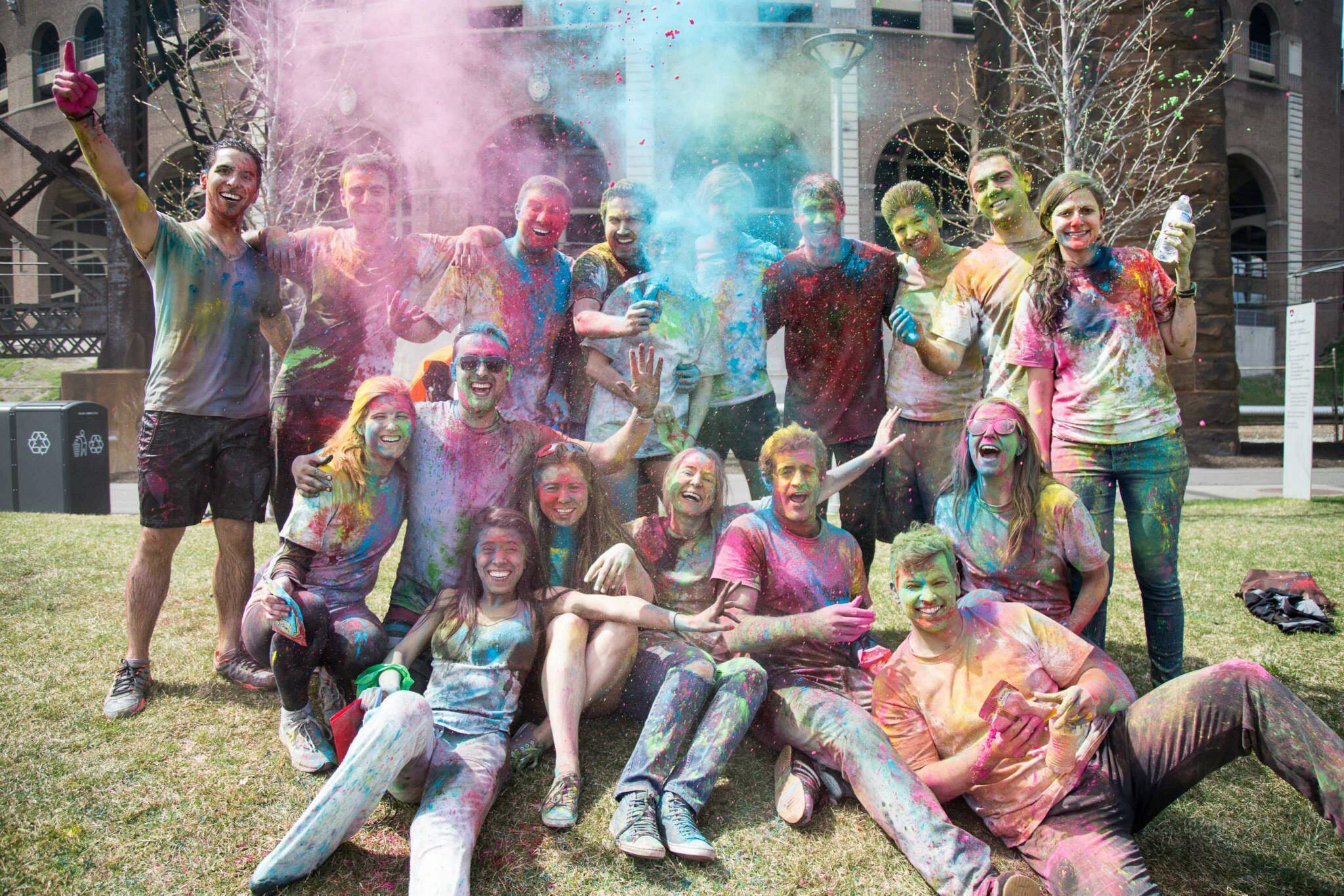 A group of people covered in vibrant colored powders, celebrating an outdoor festival in a park-like setting, possibly Holi.