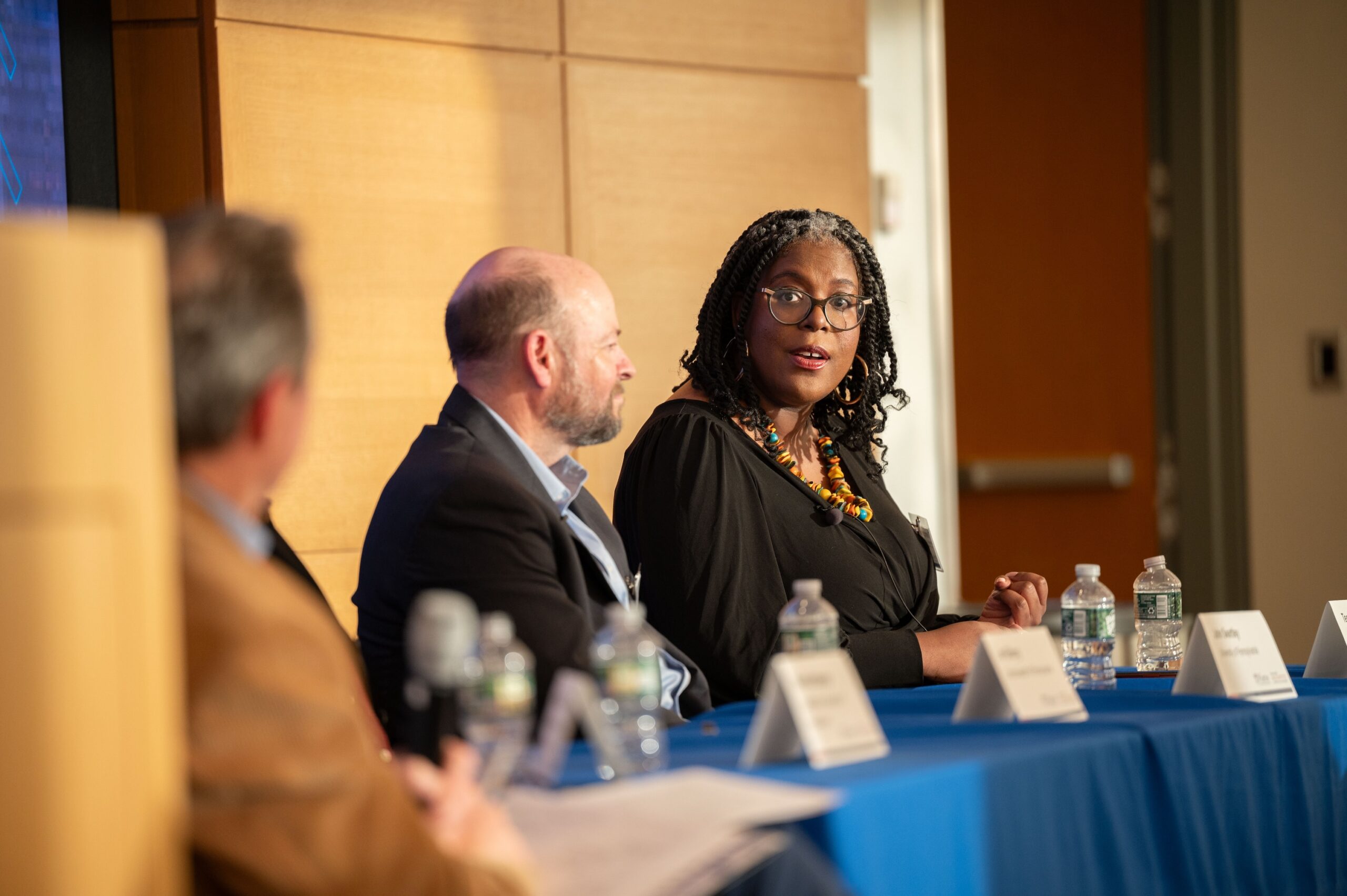 A panel discussion with several people seated at a table with name cards and water bottles. One panelist, wearing glasses and a necklace, is speaking and engaging with the audience.