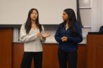 Two individuals in a classroom setting are standing in front of a projector screen, engaged in conversation or presentation.