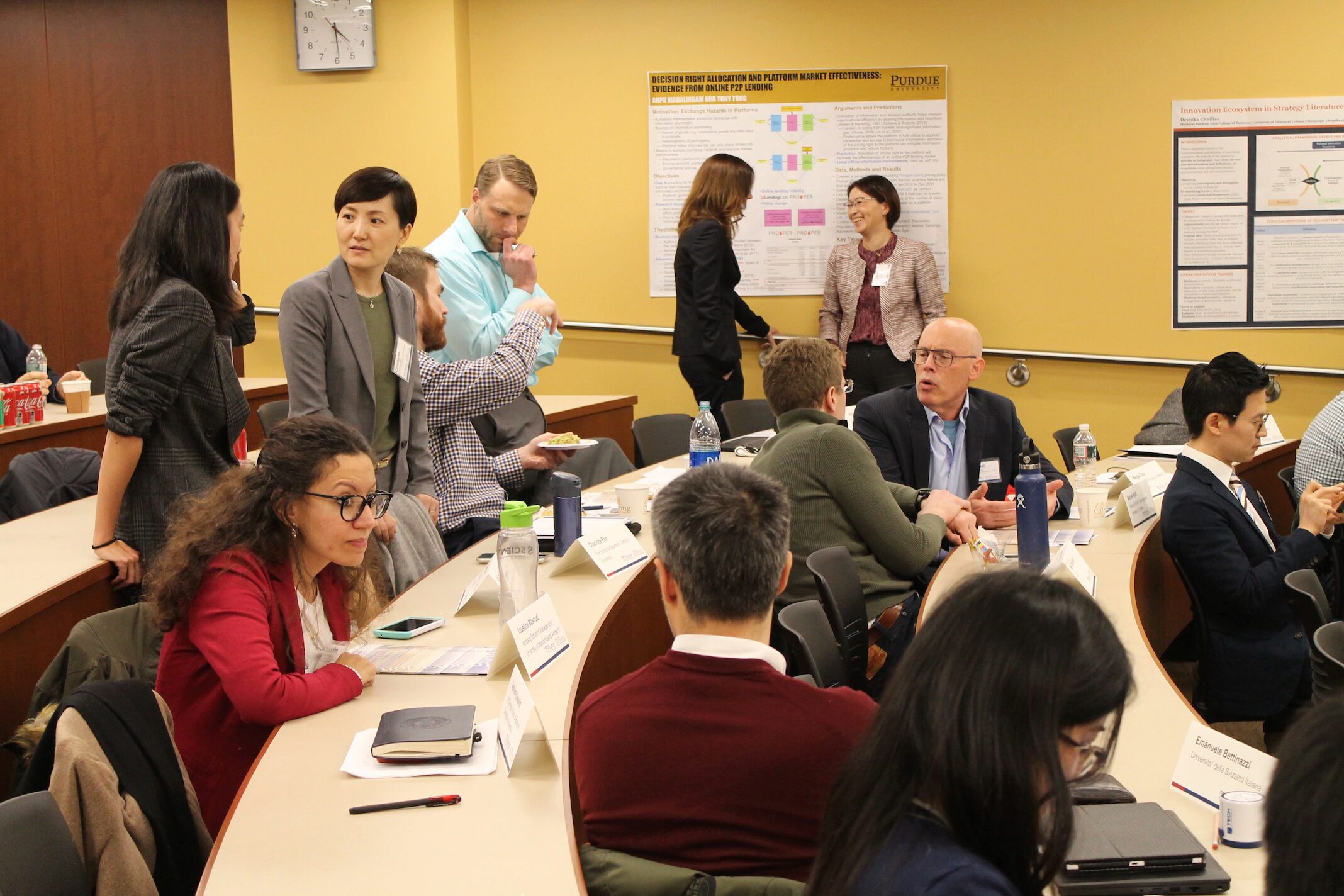 A group of people engaged in discussions in a conference or workshop setting. The room has posters on the walls and individuals seated around tables with nameplates and notes.