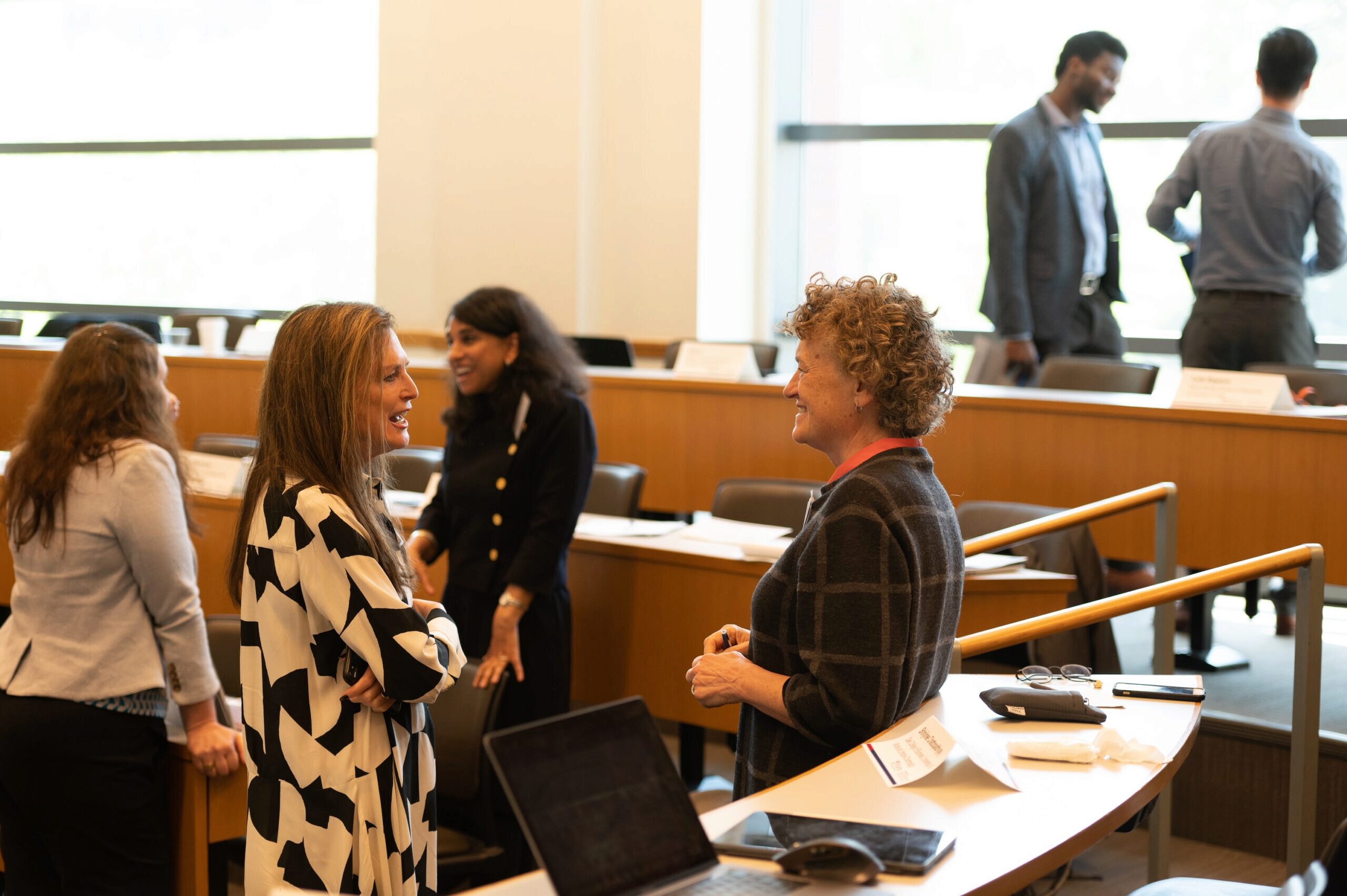 A small group of people engaged in conversation in a conference room or lecture setting. They are standing near desks with papers and electronic devices.