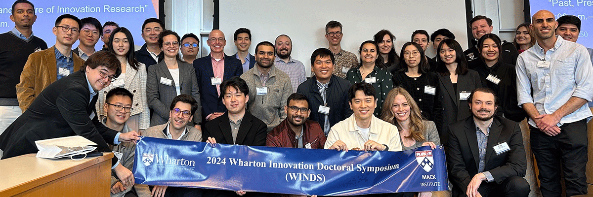 A large group of people posing together, holding a banner that reads "2024 Wharton Innovation Doctoral Symposium (WINDS)." They are in a professional setting, suggesting participation in an academic or business event.