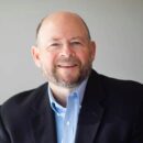 A headshot of a person with a beard and balding hair, wearing a blue button-up shirt and dark blazer, smiling against a plain background.