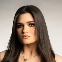 Headshot of a person with long, straight hair, wearing a neutral expression and a necklace with a circular pendant. The background is a gradient of gray tones.