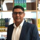 A person wearing glasses and a suit stands in front of a bookshelf filled with colorful books.