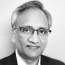 A grayscale headshot of a person with short gray hair, wearing glasses and a suit jacket, looking at the camera with a slight smile.