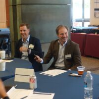 A small group of people are smiling and chatting at a round table in a conference or meeting setting. There are papers, drinks, and name tags visible.