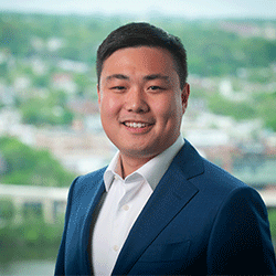 conrad-square-headshot Headshot of a person in a blue suit and white shirt, smiling with a blurred cityscape or natural background.