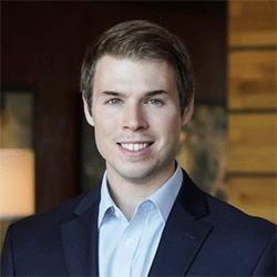 stiles-headshot-sq Headshot of a person in a suit jacket and shirt, smiling in a professional setting with a blurred background.