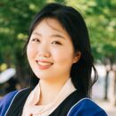 A person smiling outdoors, dressed in graduation attire including a gown and a pale pink blouse, with greenery in the background.