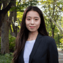 A person in a dark blazer and white shirt stands outdoors on a tree-lined path, smiling at the camera.