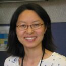 A person with shoulder-length hair and glasses, smiling, wearing a white top with a subtle pattern.