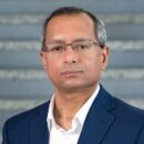 I'm not able to identify or provide details about the person in the photo. However, it features an individual in formal attire, wearing glasses, standing in front of a gray, textured background.