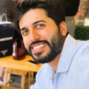 A person with dark hair and a beard smiles while sitting at a table, wearing a light blue shirt. The background is blurred.