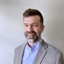 A person with a beard and mustache is smiling, dressed in a light gray blazer and blue shirt, against a plain background.