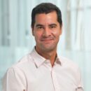 A person with short dark hair wearing a light pink collared shirt, smiling in front of a blurred light background.