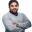 A person with short, dark hair and a beard, wearing a light gray sweater and a wristwatch, standing with arms crossed against a white background.