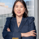 A person with dark hair wearing a denim jacket, arms crossed, standing indoors near a window with a blurred cityscape background.