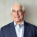 A person wearing a blue suit and light blue shirt smiles while posing against a plain background.