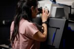 Person in a pink shirt using a microscope in a laboratory setting.