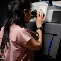 Person in a pink shirt using a microscope in a laboratory setting.