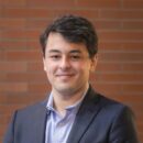 A person in a suit and shirt posing in front of a brick wall, smiling slightly.