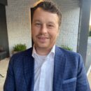 A person in a blue checked suit jacket and white shirt stands in front of a white brick wall, smiling.